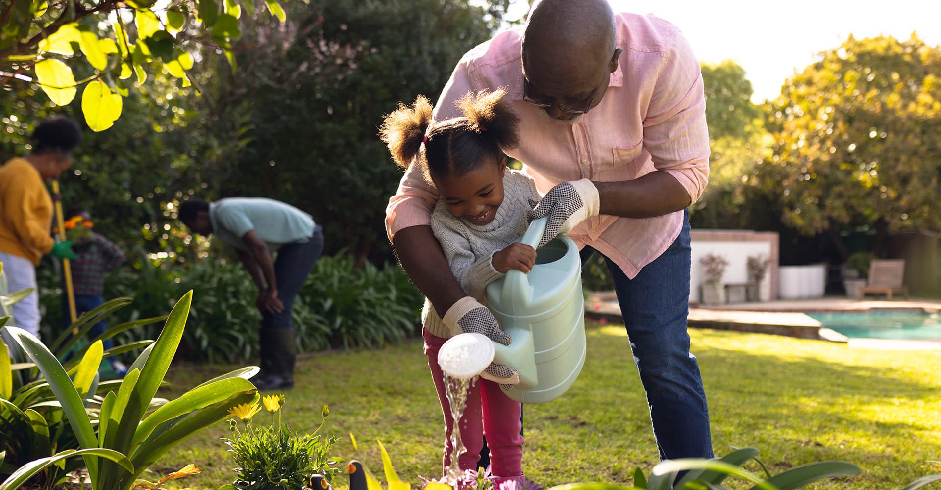 A grandfather and granddaughter watering flowers in a garden bed in their yard
