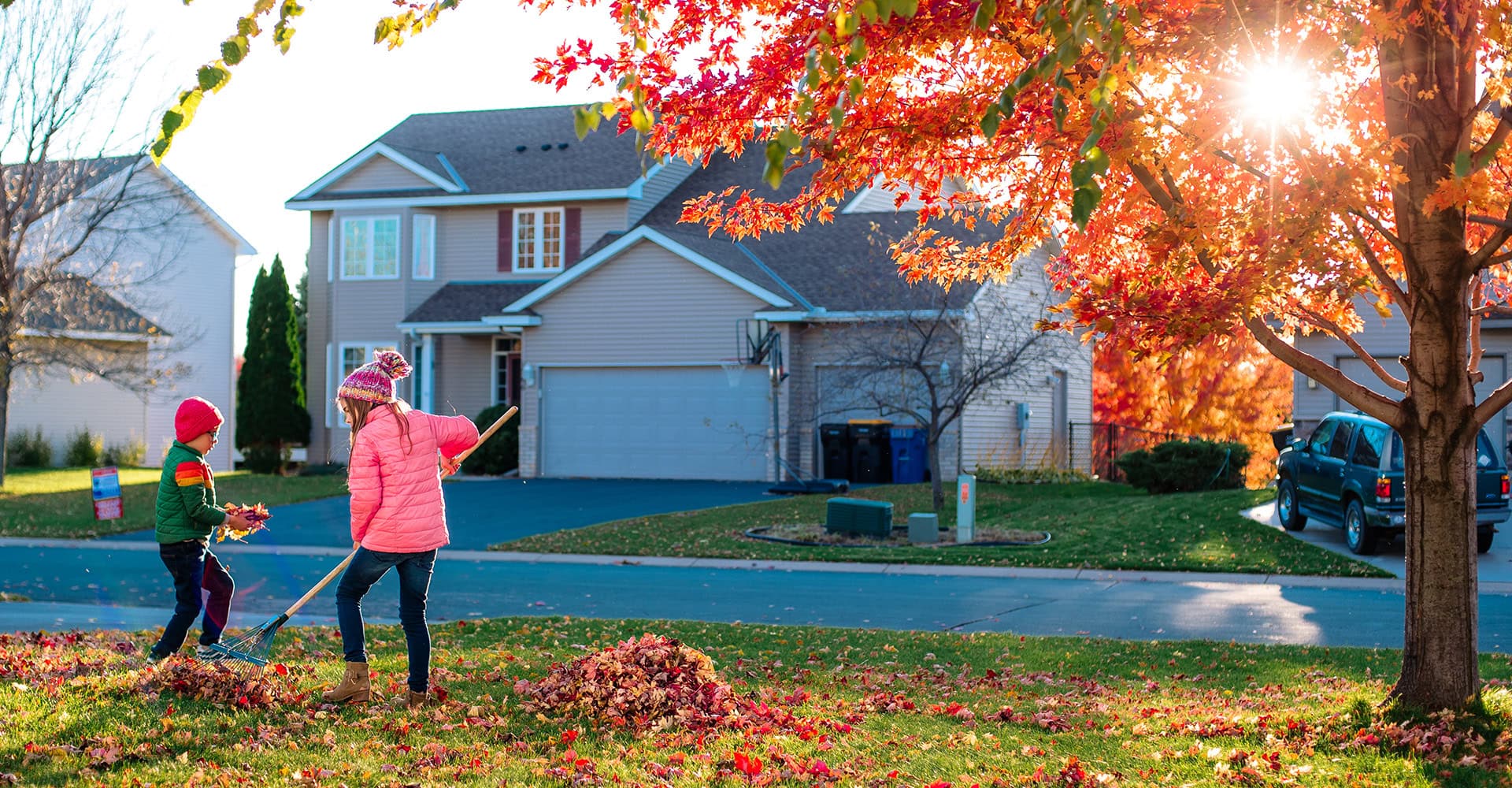 Two children wearing coats raking orange leaves in a front yard.